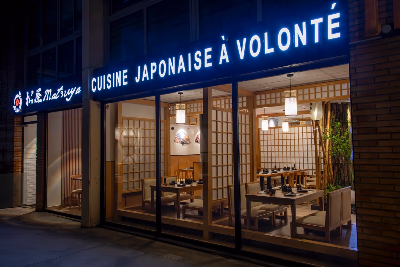 Façade extérieure d'un restaurant japonais nommé Matsuya avec un panneau lumineux indiquant 'CUISINE JAPONAISE À VOLONTÉ' et vue sur l'intérieur moderne avec tables et chaises en bois.