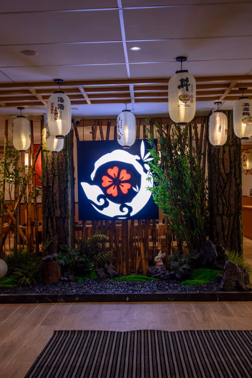 Entrée décorée d'un restaurant japonais avec des lanternes suspendues, un panneau lumineux avec un motif floral rouge, des plantes et un lapin en décoration.