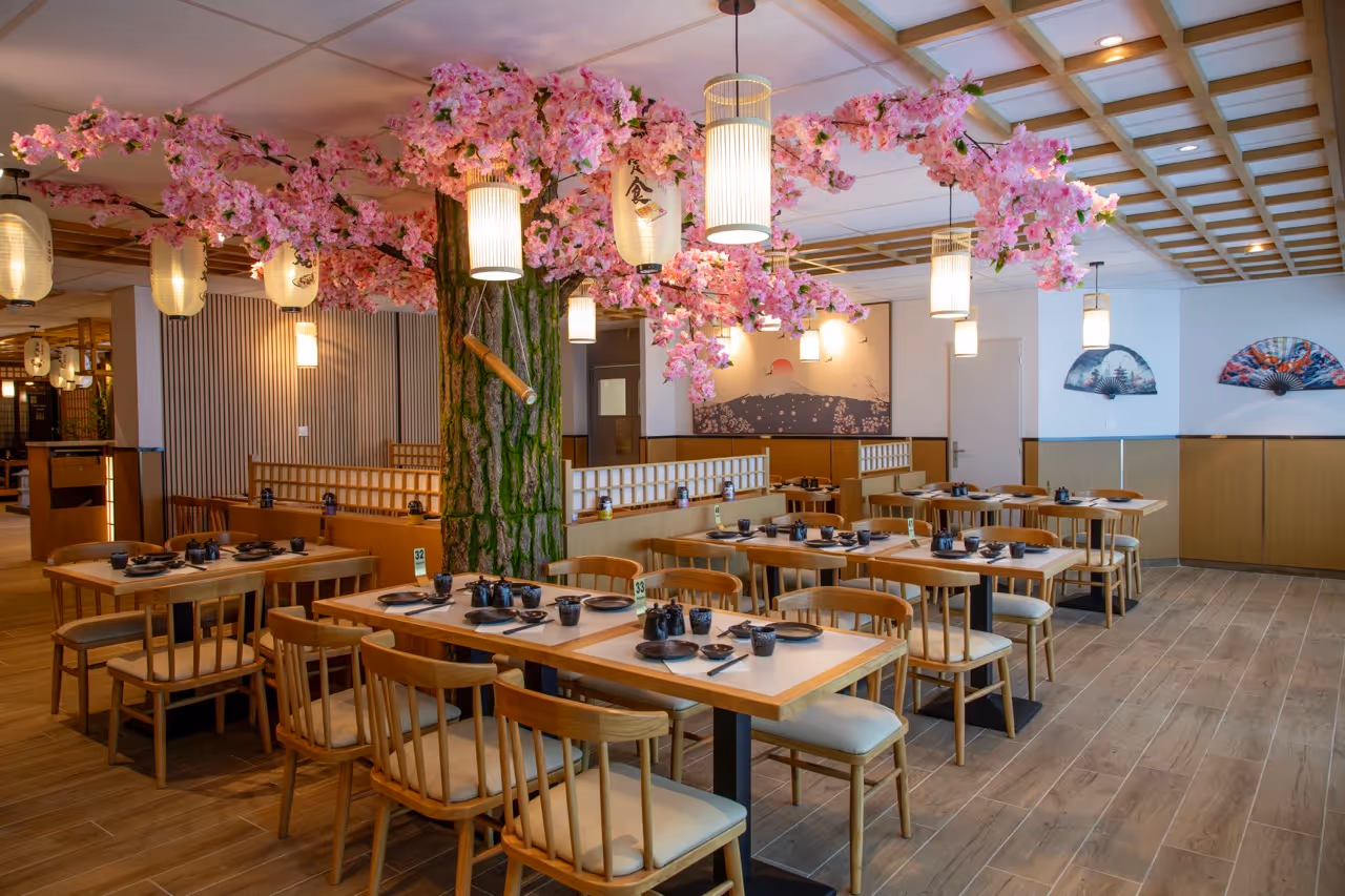 Intérieur d'un restaurant japonais avec une grande fausse cerisier en fleurs au centre et des tables en bois disposées autour.