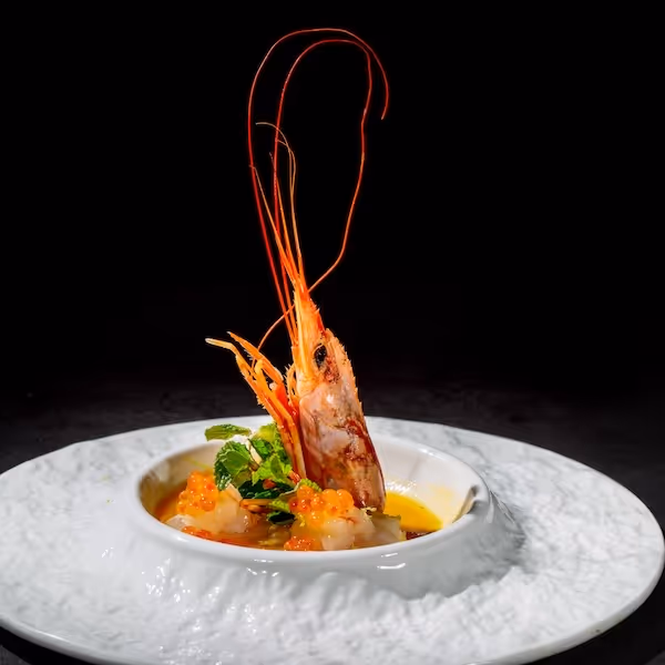 Plat gastronomique avec une crevette entière dressée au centre, sur un lit de morceaux de poisson cru, œufs de poisson orange et une feuille de menthe dans une assiette blanche texturée.
