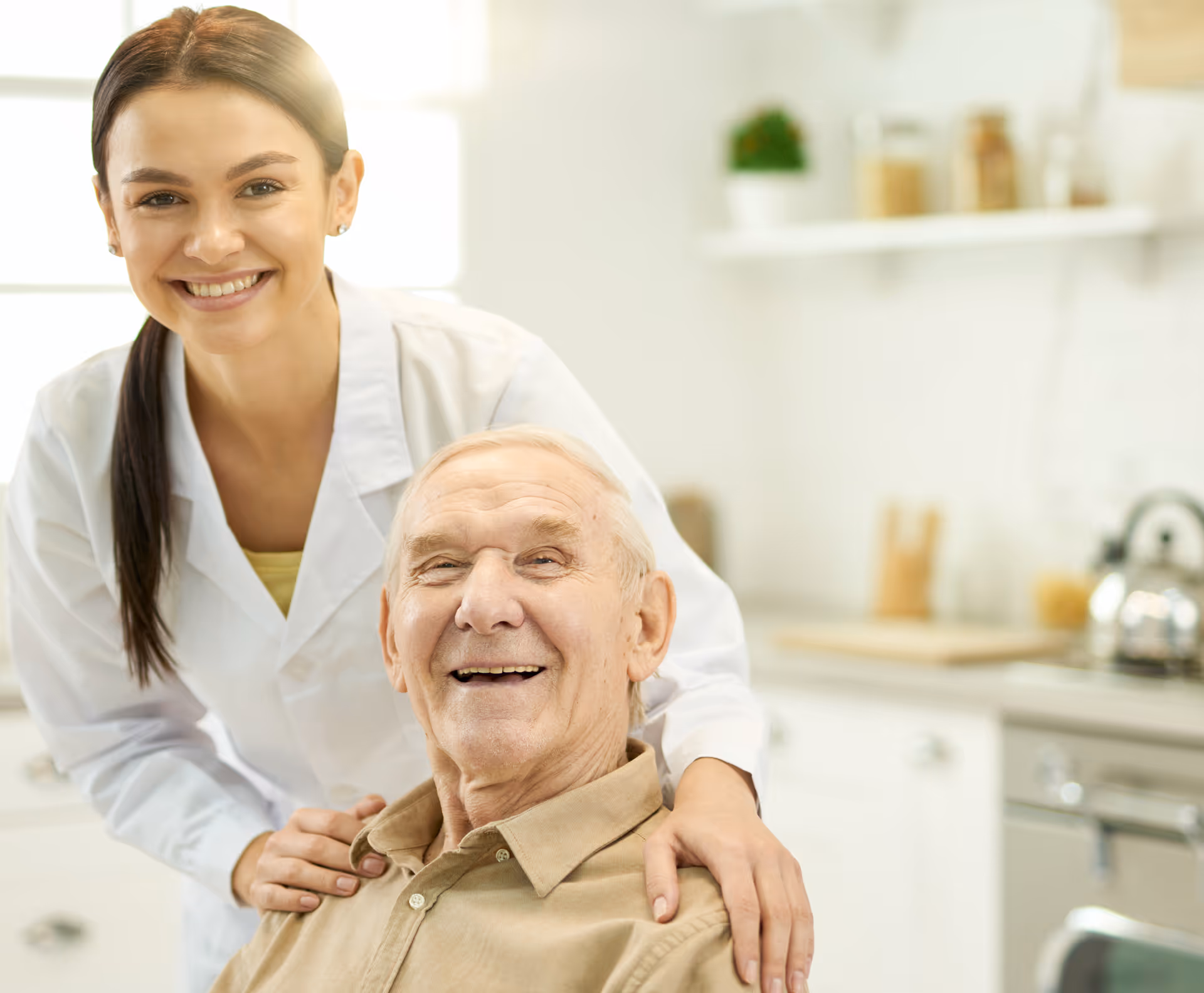 Eine weibliche Pflegefachkraft umarmt zierlich den Klienten im Rollstuhl an der Schulter und beide lächeln dabei in die Kamera.