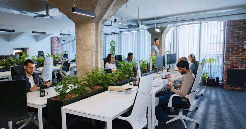 Modern open office with white desks, plants, and people working on computers and tablets.