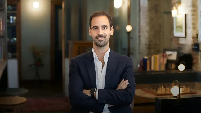 Man in a dark blazer and white shirt standing with arms crossed in a warmly lit modern office lounge.