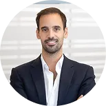 Smiling man with short brown hair wearing a dark blazer and white shirt in front of a blurred background.