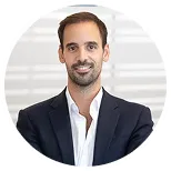 Smiling man with short dark hair and beard wearing a dark suit jacket and white shirt against a blurred background.