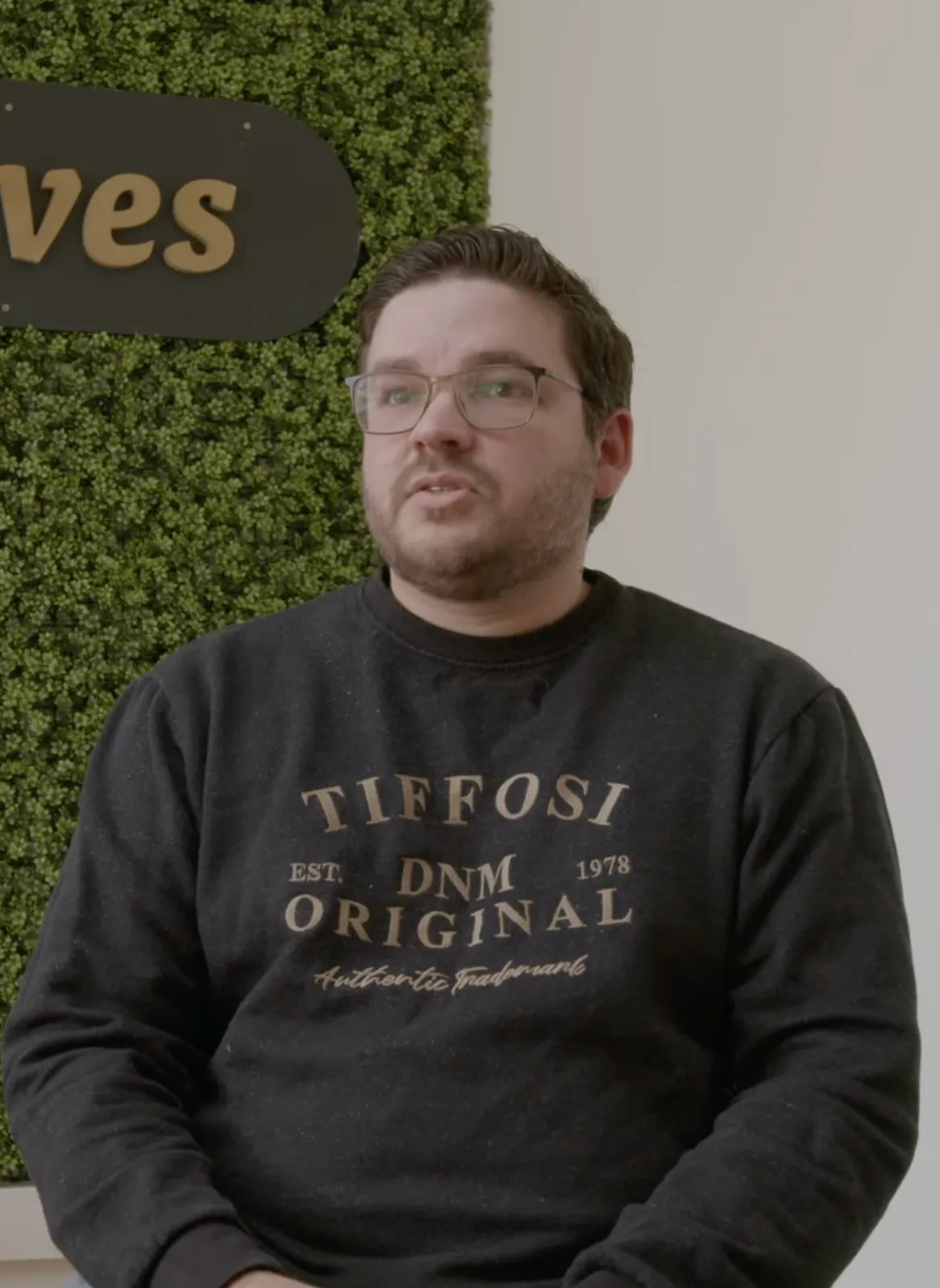 Man wearing glasses and a black sweatshirt with 'TIFFOSI DNM ORIGINAL' text, sitting against a green leafy wall and white background.