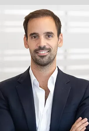 Smiling man with dark hair and beard wearing a white shirt and dark blazer, standing with arms crossed.