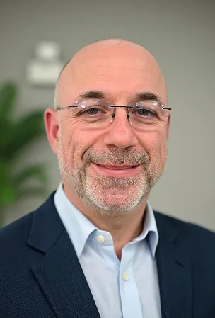 Smiling bald man with glasses and gray beard wearing a navy blazer and light blue shirt.