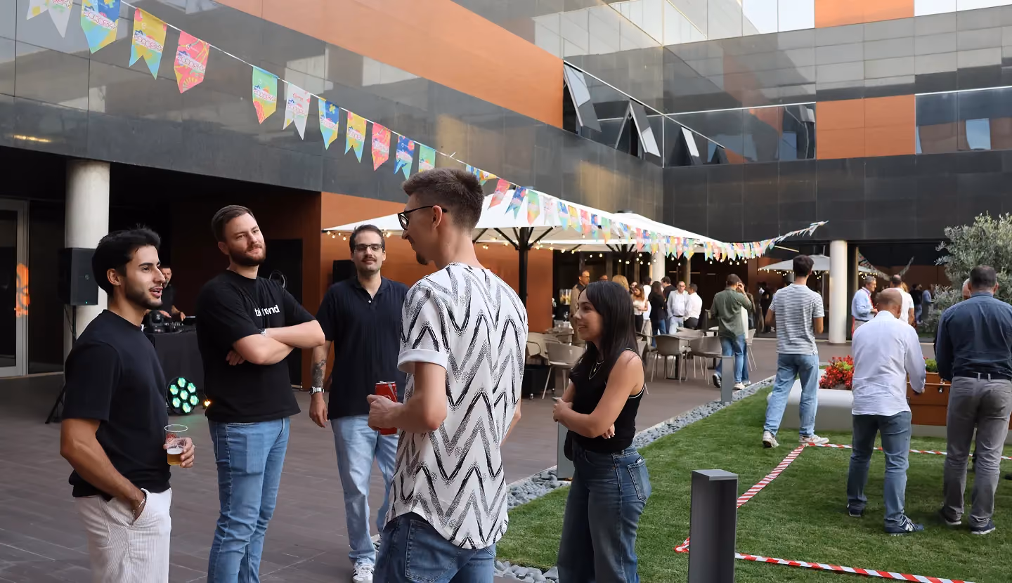 Mensen die socializen tijdens een buitenevenement met kleurrijke vlaggen boven hun hoofden en tafels op de achtergrond.