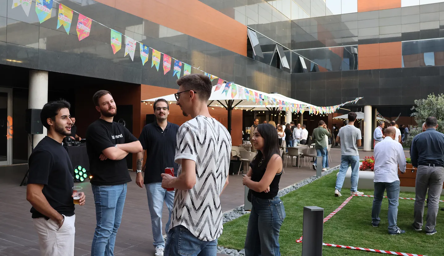 People socializing at an outdoor event with colorful flags hanging overhead and tables in the background.