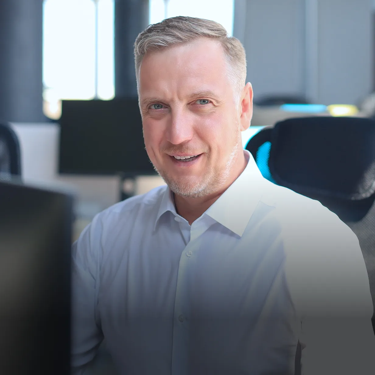 Smiling middle-aged man with short blond hair and blue eyes wearing a white shirt, sitting in an office.