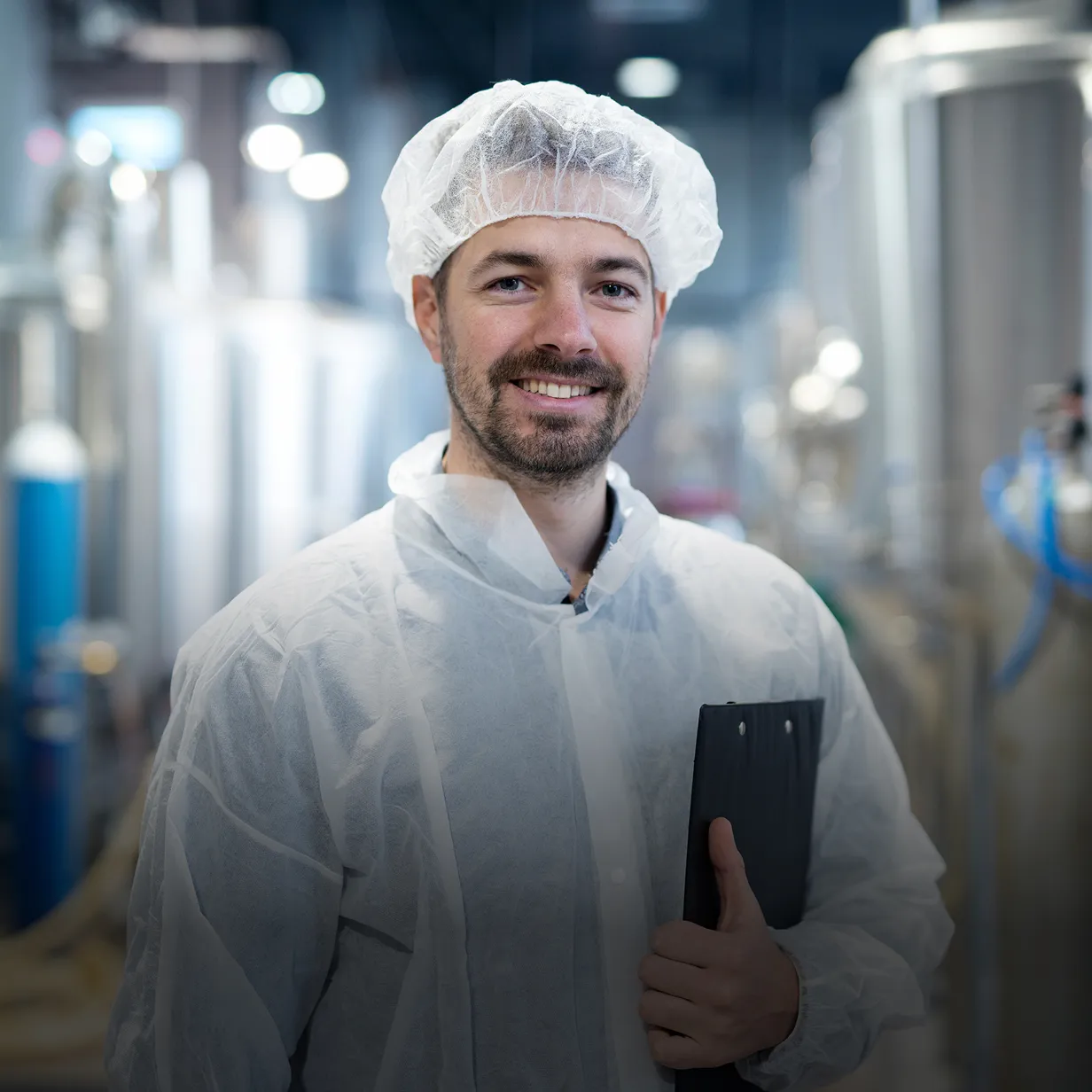 Smiling male worker in white protective suit and hairnet holding a clipboard in an industrial facility.