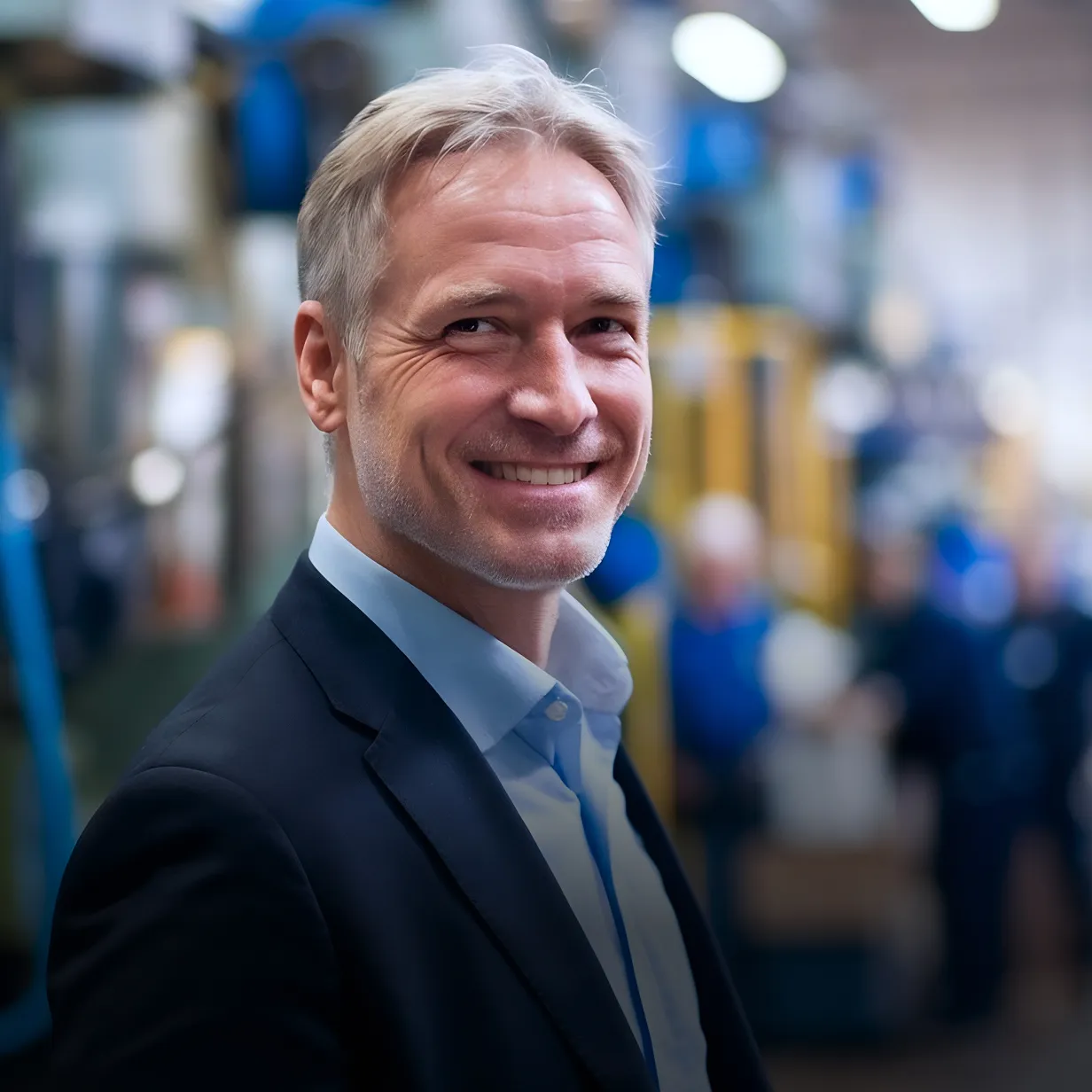 Smiling middle-aged man in a suit standing in a blurred industrial or factory setting.