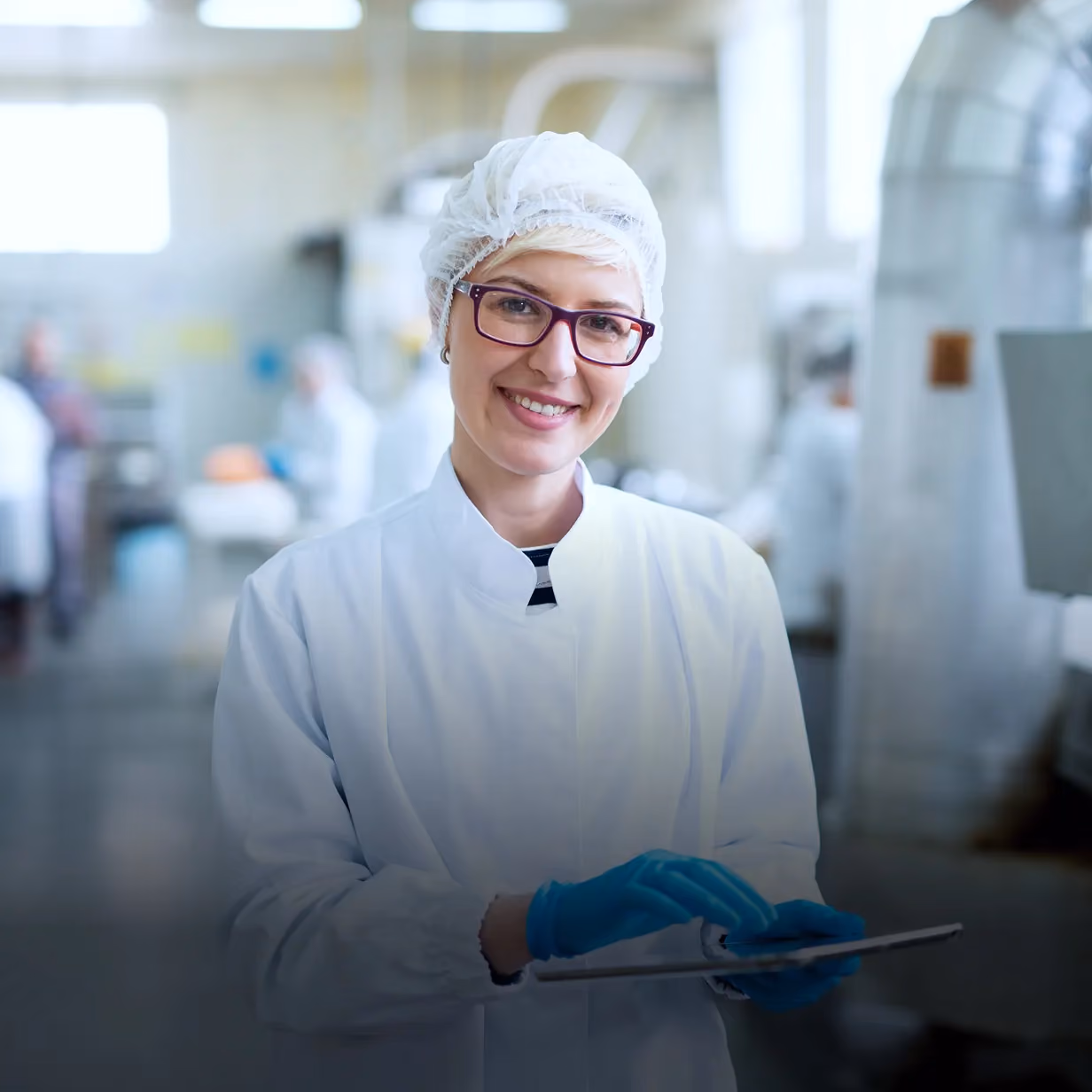 Smiling woman in white lab coat, hairnet, glasses, and blue gloves holding a tablet in a bright industrial or laboratory setting.