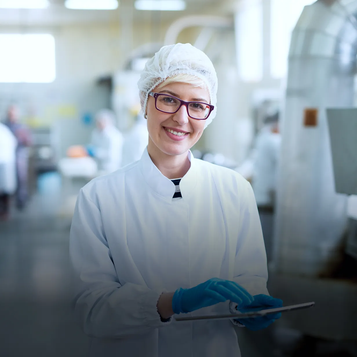 Smiling woman in white lab coat, hairnet, glasses, and blue gloves holding a tablet in a bright industrial or laboratory setting.