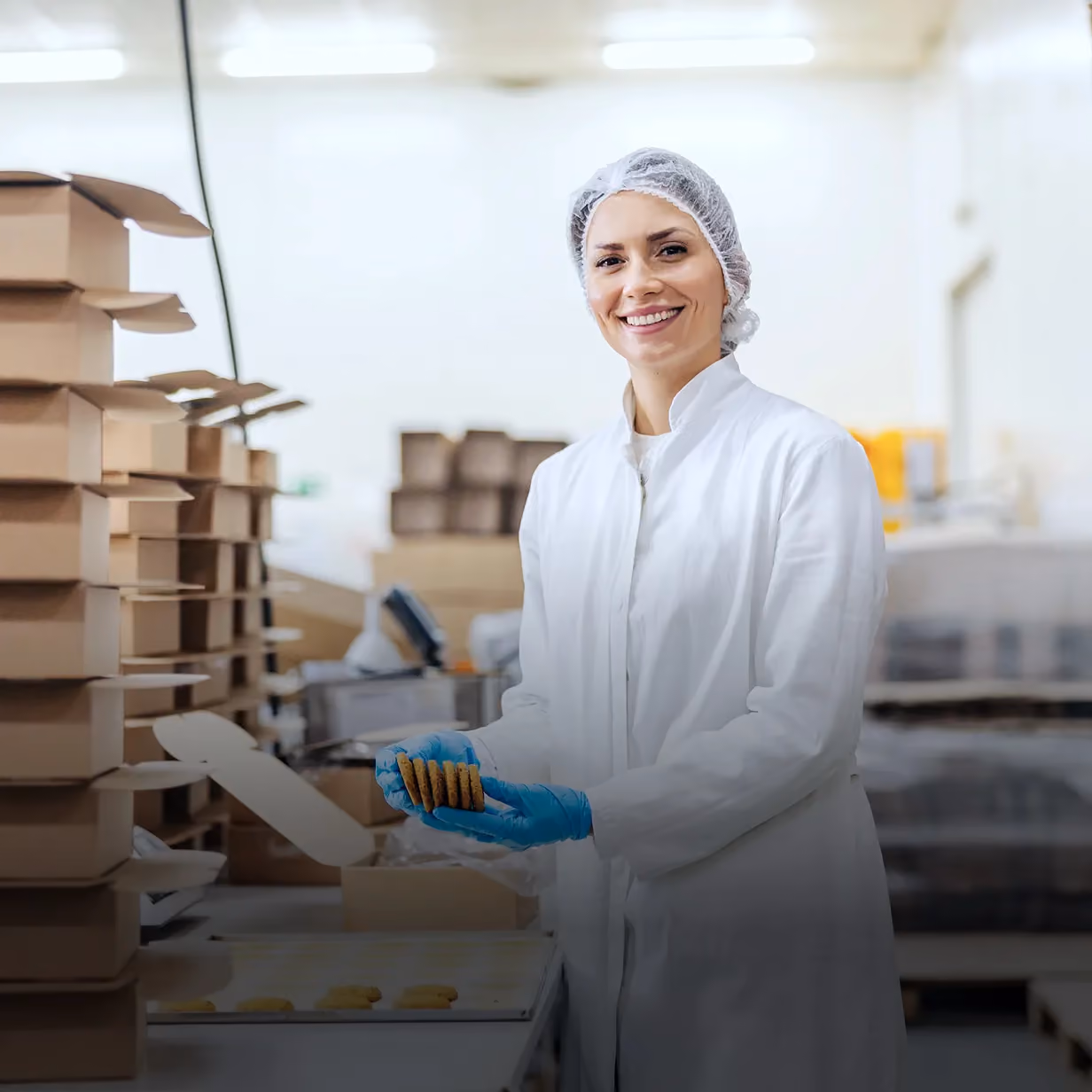 Smiling food factory worker in a white coat and hairnet holding cookies with blue gloves in a packaging area.
