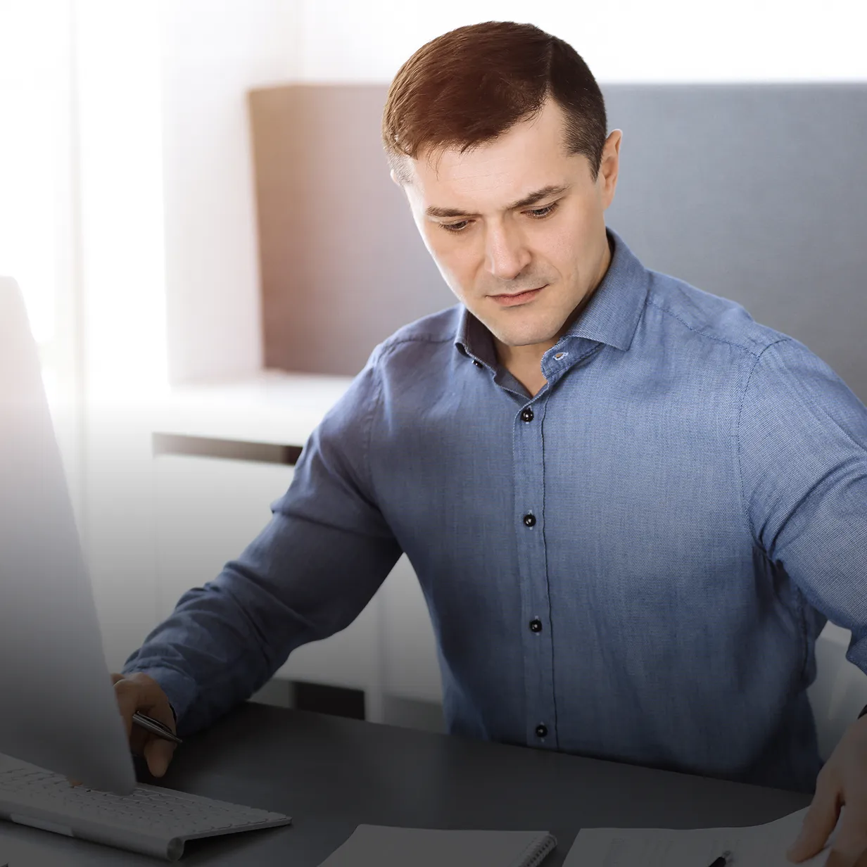 Man in a blue button-up shirt working at a desk with a computer and papers.