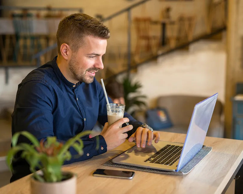 Smiling man in navy shirt drinking iced coffee while using laptop at a wooden table in a cozy cafe.