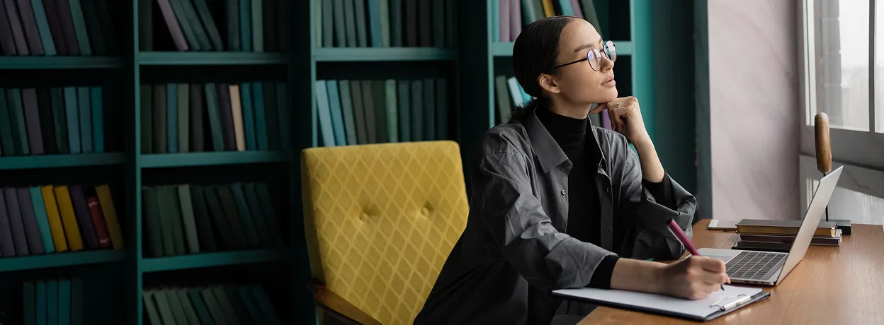 Woman wearing glasses sitting at a desk with notebook and laptop, looking thoughtfully out a window in a room with bookshelves.