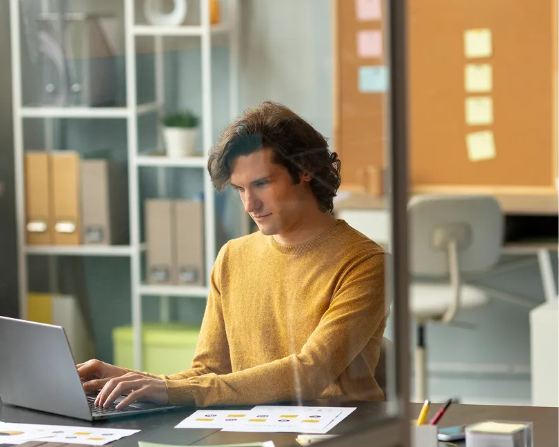 Man in a mustard sweater working on a laptop at a desk with papers and office supplies.