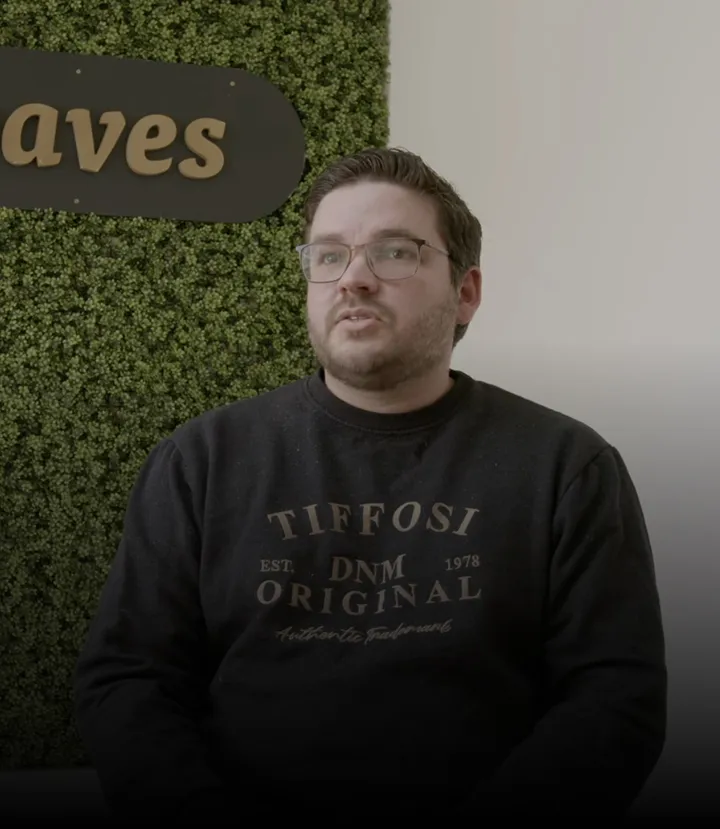 Man with glasses and beard wearing a black Tiffosi sweatshirt sitting in front of a green wall with partial 'aves' sign.