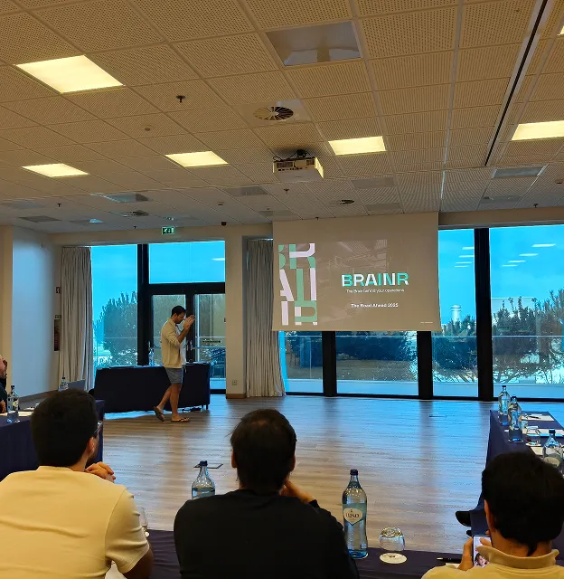 Presenter speaking in front of a screen displaying the text 'BRAINR' in a conference room with seated attendees and large windows overlooking trees and a patio.