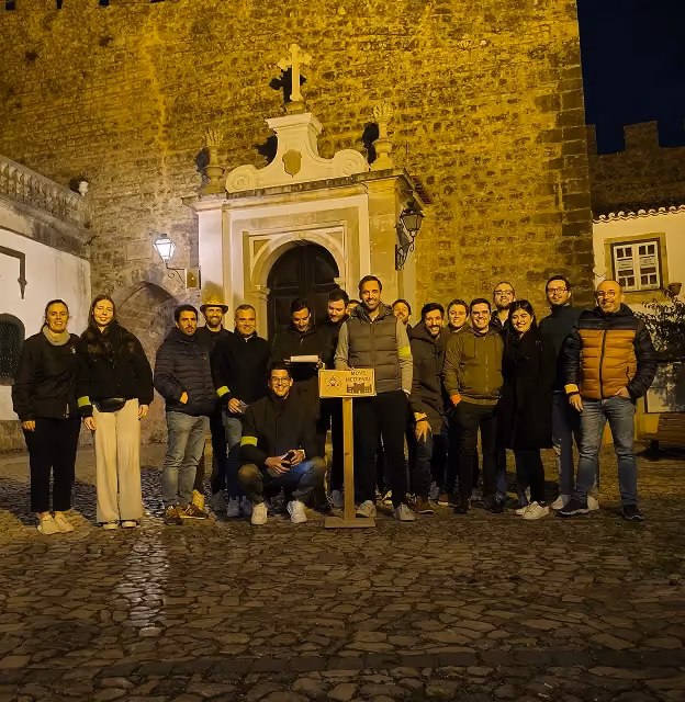 Groep mensen die 's nachts poseert voor een oud stenen gebouw met een gewelfde deuropening en een kruis bovenop.