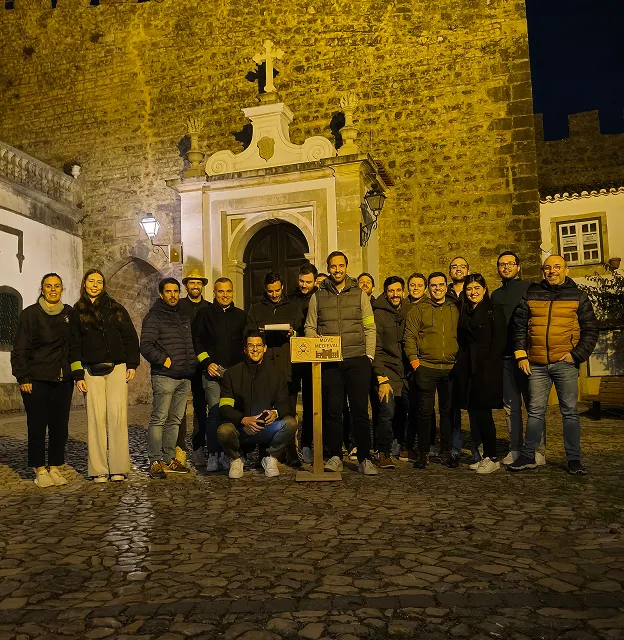Group of people posing at night in front of an old stone building with an arched doorway and a cross on top.