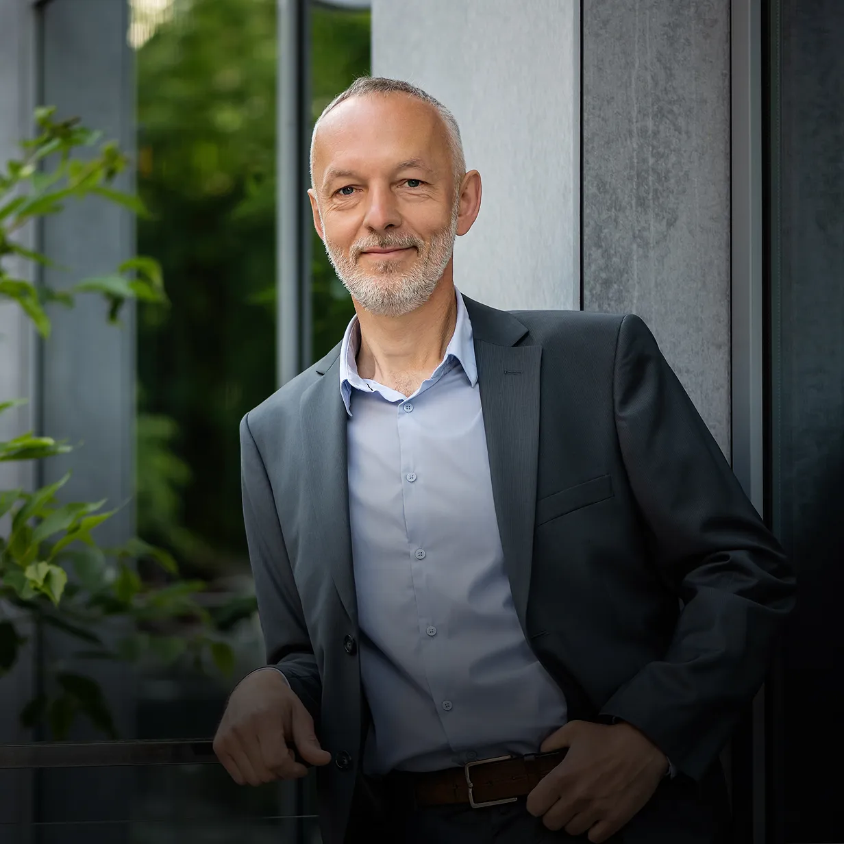 Smiling middle-aged man with grey beard in a dark suit leaning against a window with greenery in the background.