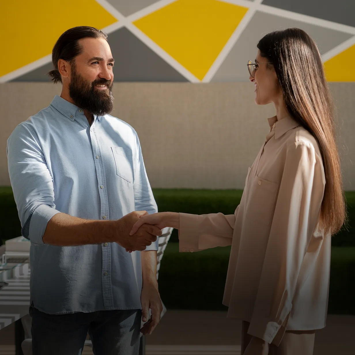 Man with beard and blue shirt shaking hands with woman wearing glasses and beige blouse in a modern office setting.