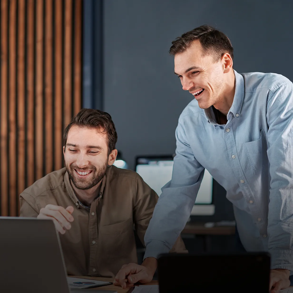 Two men smiling and looking at a laptop screen in an office setting.