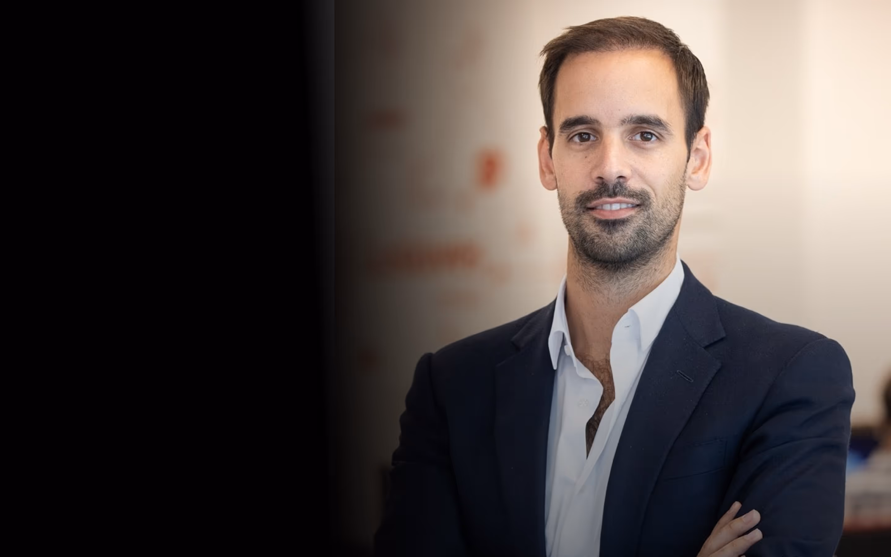 Smiling man in a navy suit with arms crossed sitting in a modern office setting.