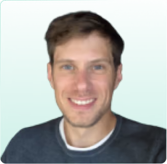 Homem sorridente, com cabelo castanho curto e olhos azuis, vestindo uma camisa cinza-escura sobre um fundo verde.