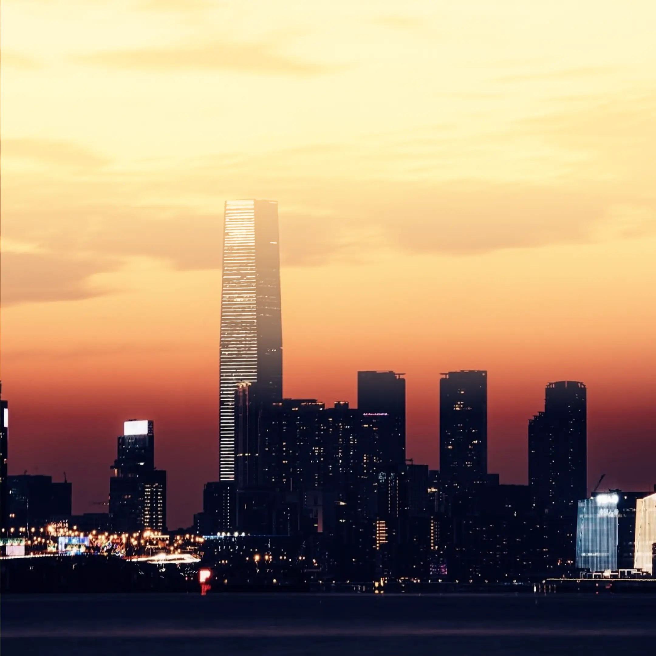City skyline at dusk with a tall illuminated skyscraper and an orange-yellow sunset sky.