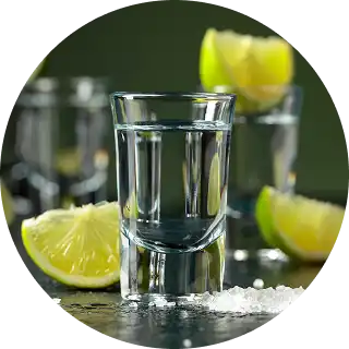Clear shot glass filled with liquid, surrounded by lemon wedges and salt on a reflective surface.