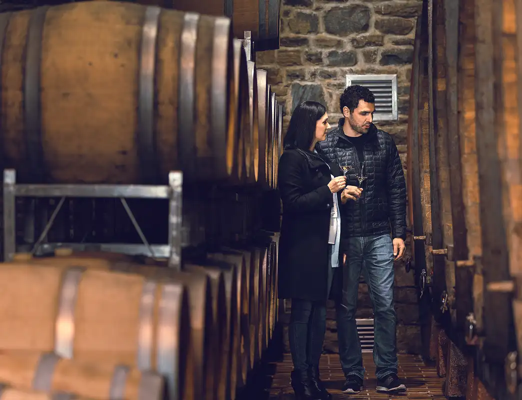 Man and woman holding wine glasses standing between rows of wooden barrels in a stone-walled cellar.