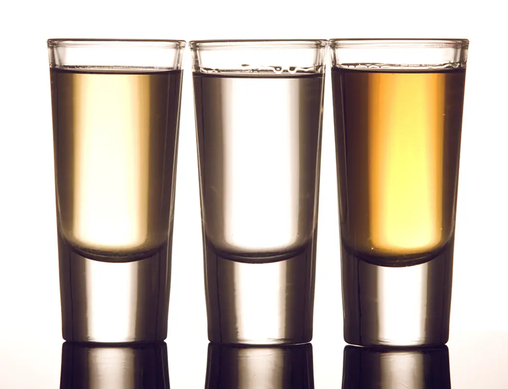 Three shot glasses filled with clear and amber-colored liquids on a reflective surface.