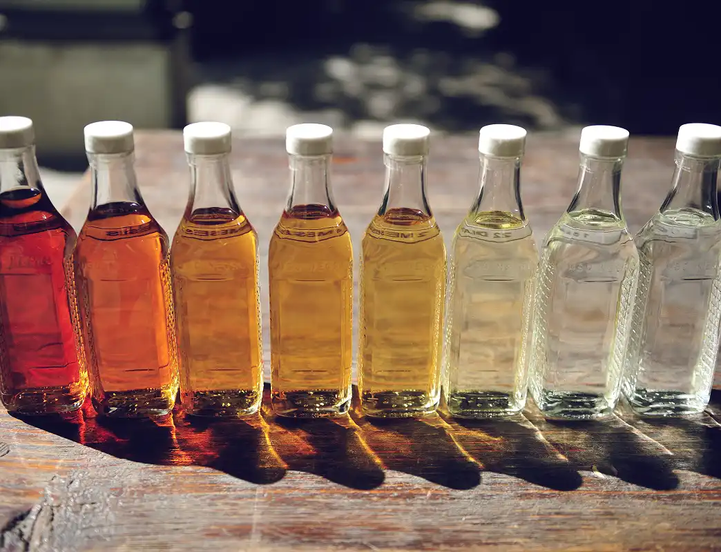 Nine small glass bottles lined up on a wooden surface, each filled with liquid ranging in color from dark amber to clear.
