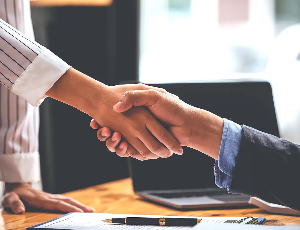 Two people shaking hands over a desk with a pen and laptop in the background.