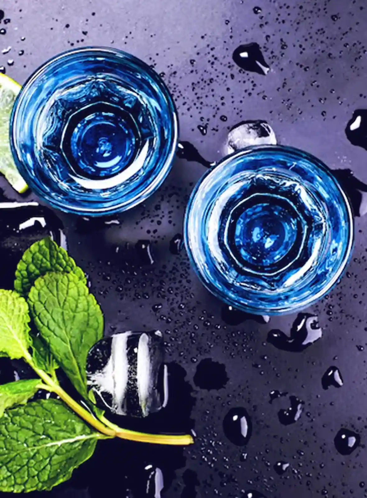 Overhead view of two blue glasses on a wet dark surface with ice cubes, fresh mint leaves, and a lemon wedge.