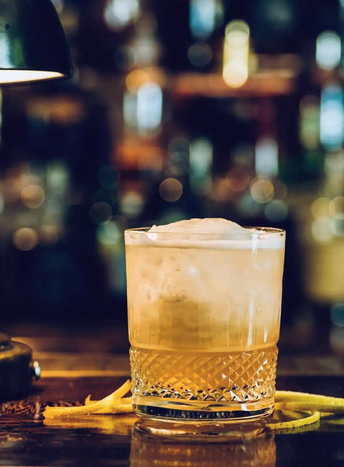 Whiskey cocktail with foam in a textured glass on a bar counter with lemon peel garnish.