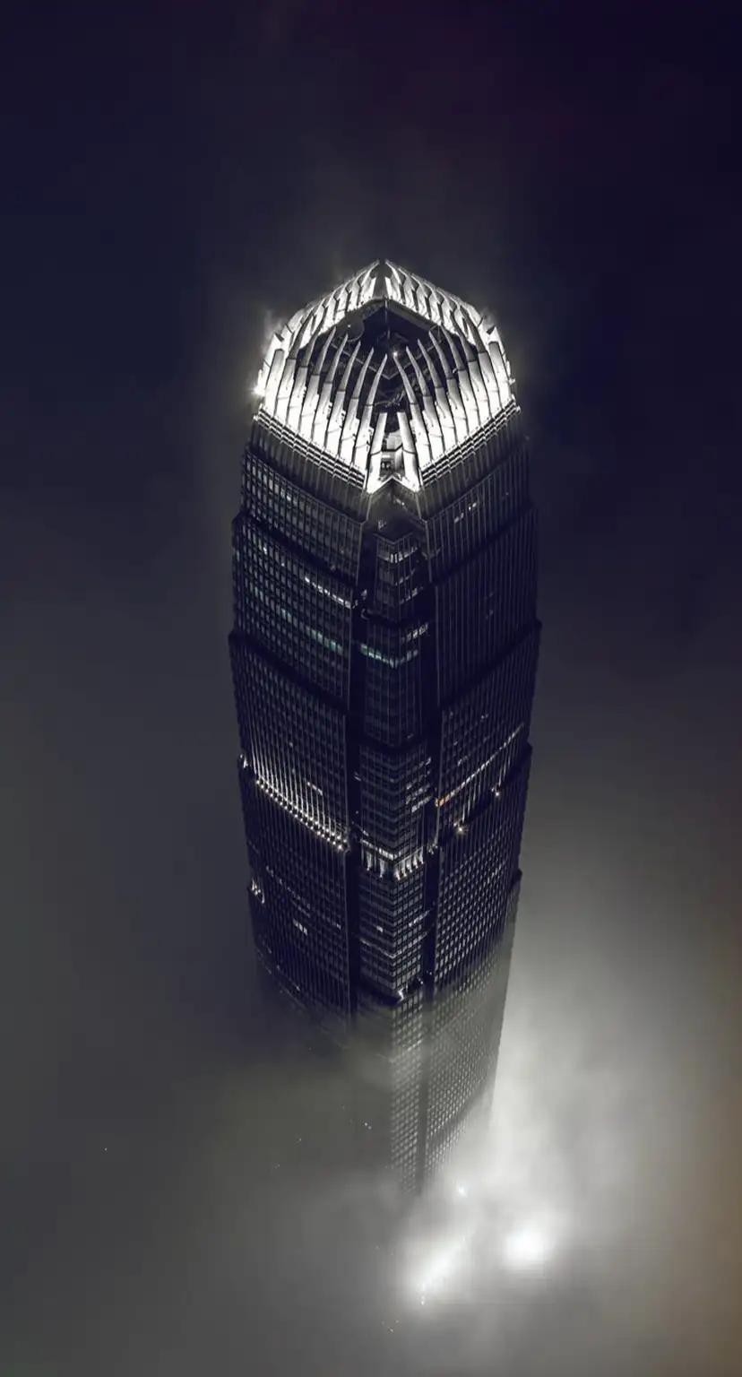 Tall skyscraper illuminated at night, rising above thick fog with bright lights glowing at the top and base.