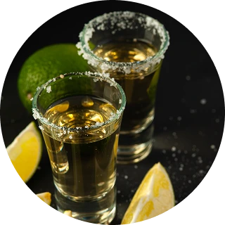 Two salt-rimmed shot glasses filled with golden tequila, accompanied by lime wedges on a dark surface.