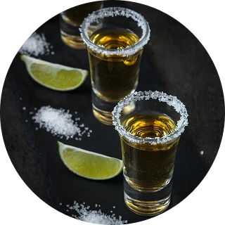 Two shot glasses of golden tequila rimmed with salt beside lime wedges and scattered salt on a dark surface.