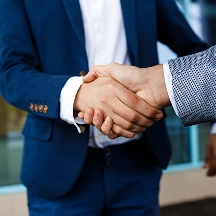 Close-up of two men shaking hands, one wearing a blue suit and the other wearing a checkered blazer.