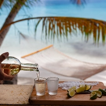 Hand pouring a drink into a glass on a wooden table with lime wedges, palm leaves, and a hammock by the beach in the background.