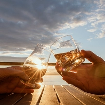 Two hands clinking glasses of whiskey together at sunset over a wooden deck by the water.