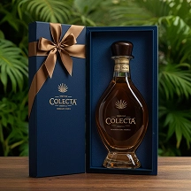 Dark glass bottle of Colecta tequila in a blue gift box with a gold ribbon, set against a green leafy background.