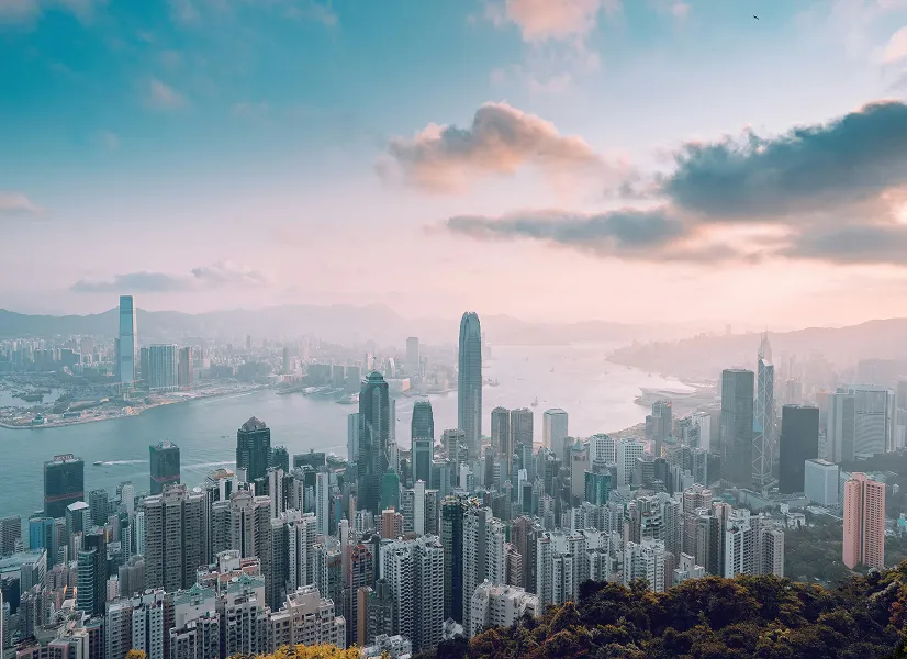 Panoramic view of a dense city skyline with skyscrapers along a harbor under a partly cloudy sky at sunset.
