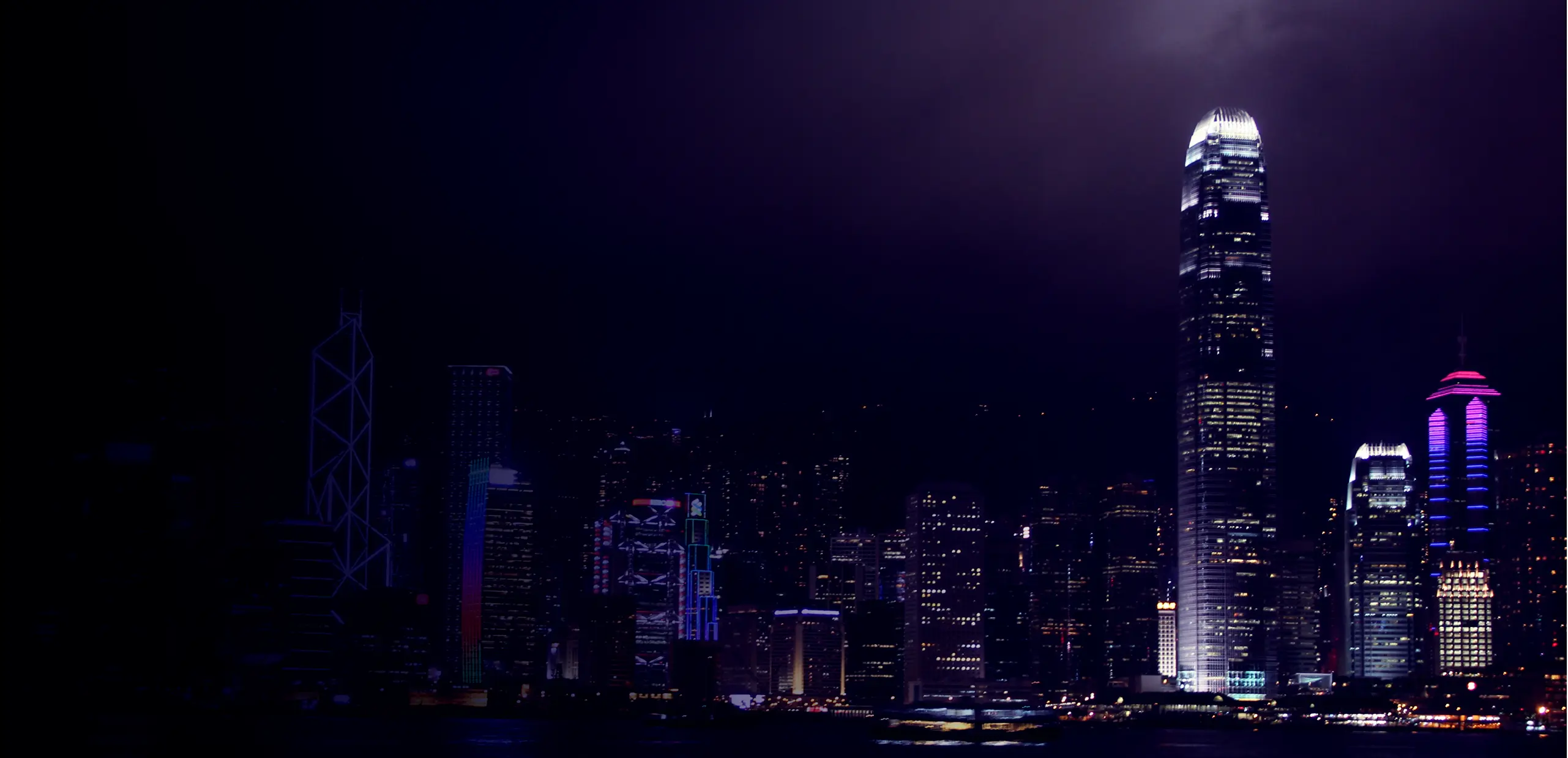 Nighttime city skyline with illuminated skyscrapers reflecting on water, featuring tall modern buildings with colorful lights.